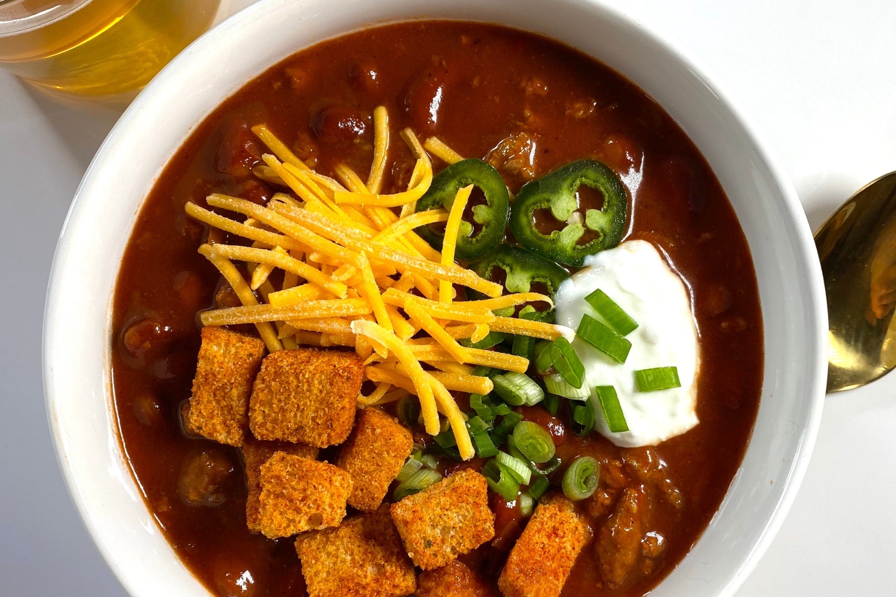 a bowl of chili with croutons, cheese, sour cream and jalapenos with a beer placed next to the bowl.