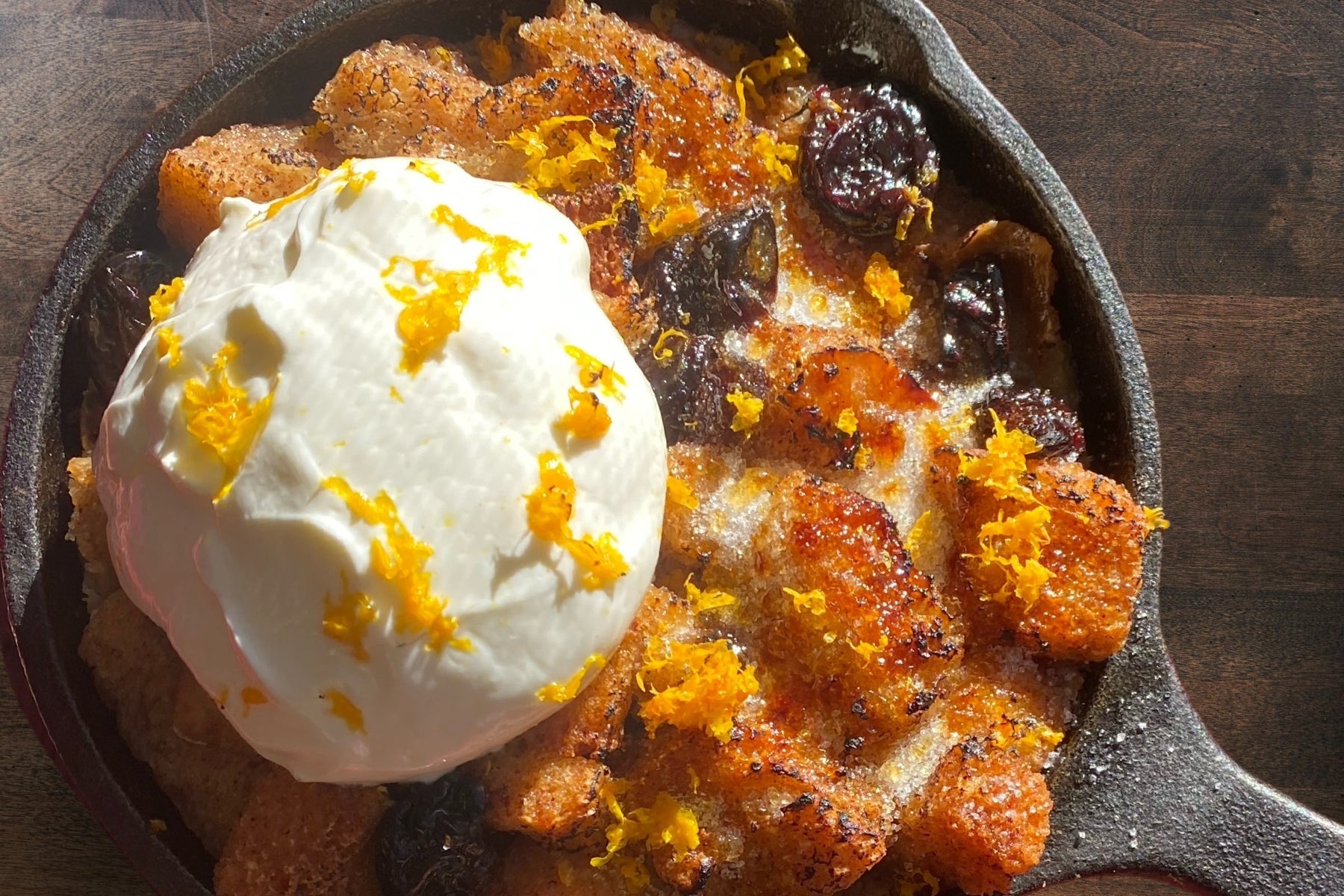 a bread pudding in a black cast iron pan with a scoop of whipped cream and orange zest.