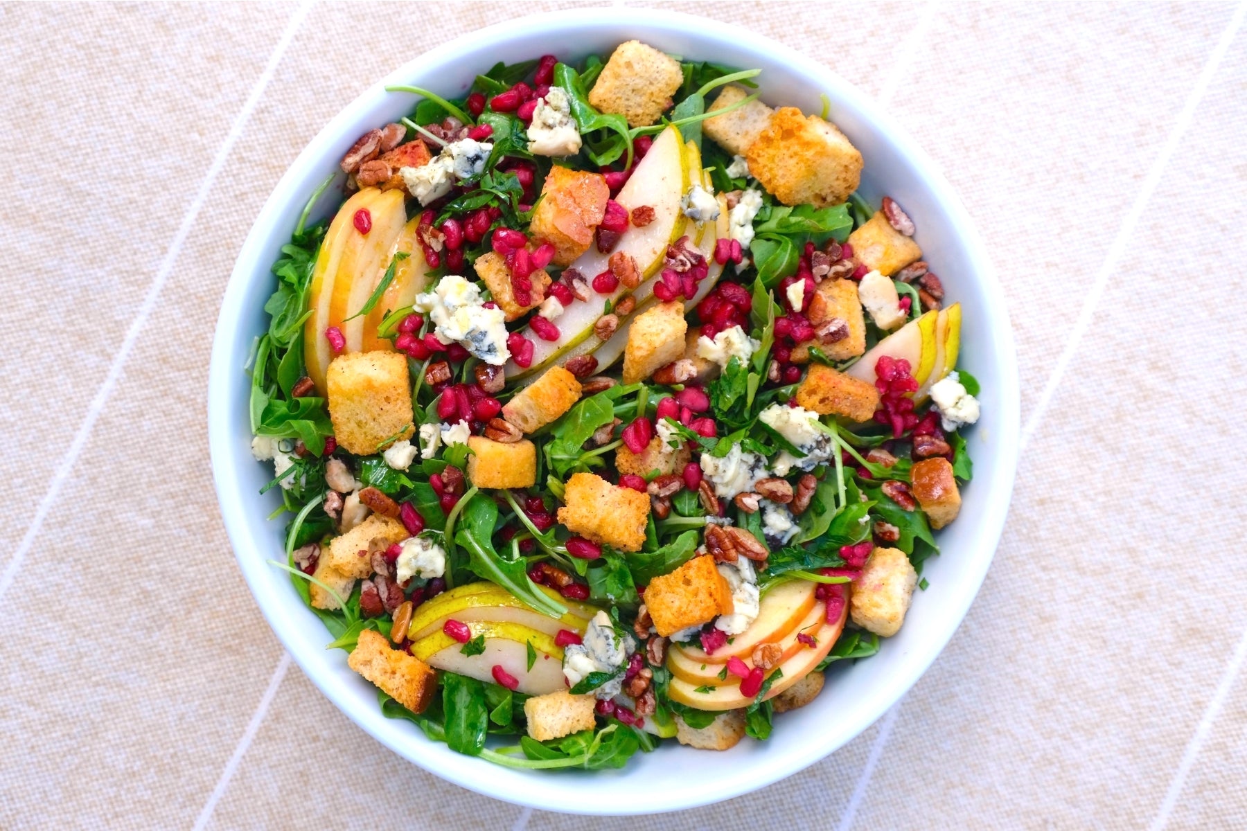 a salad bowl with colorful foods like pomegranates, pears, croutons, feta cheese and arugula.