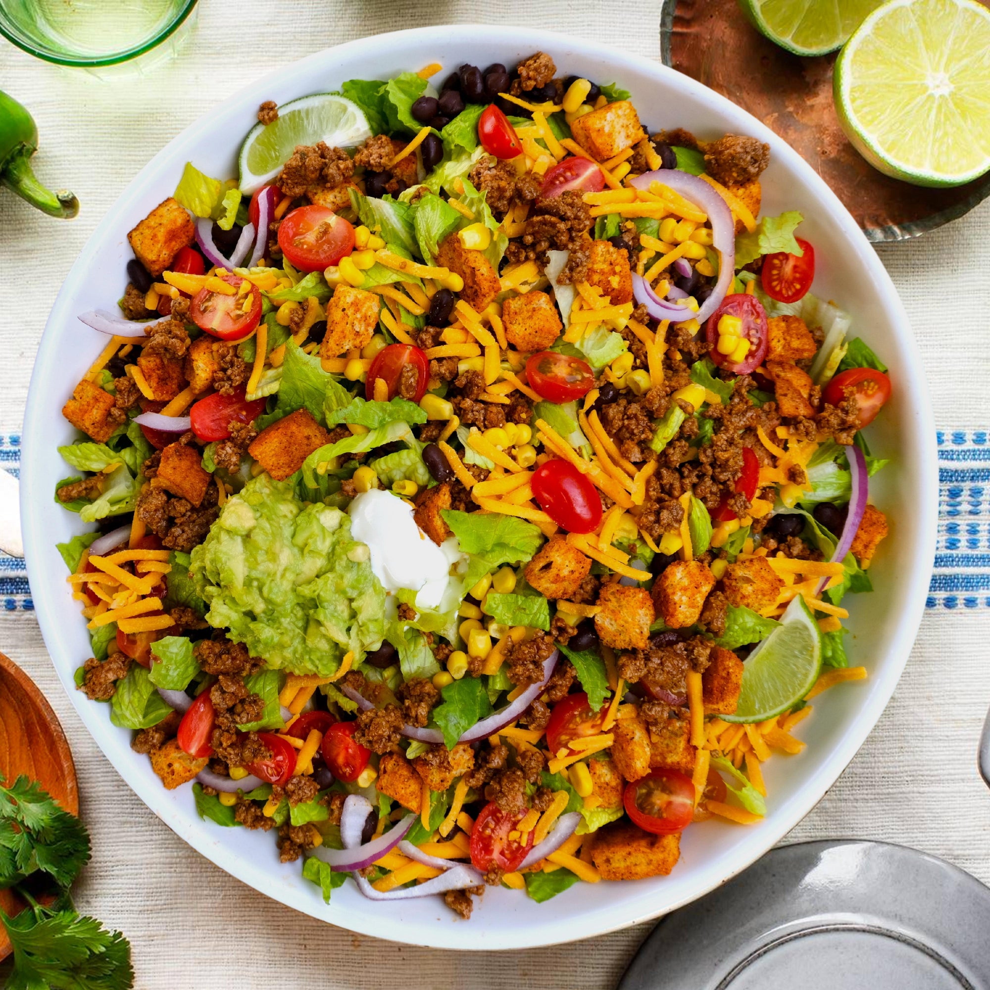 Bowl of taco salad with croutons, avocado, cheese, taco meat, tomatoes and limes on a festive table.