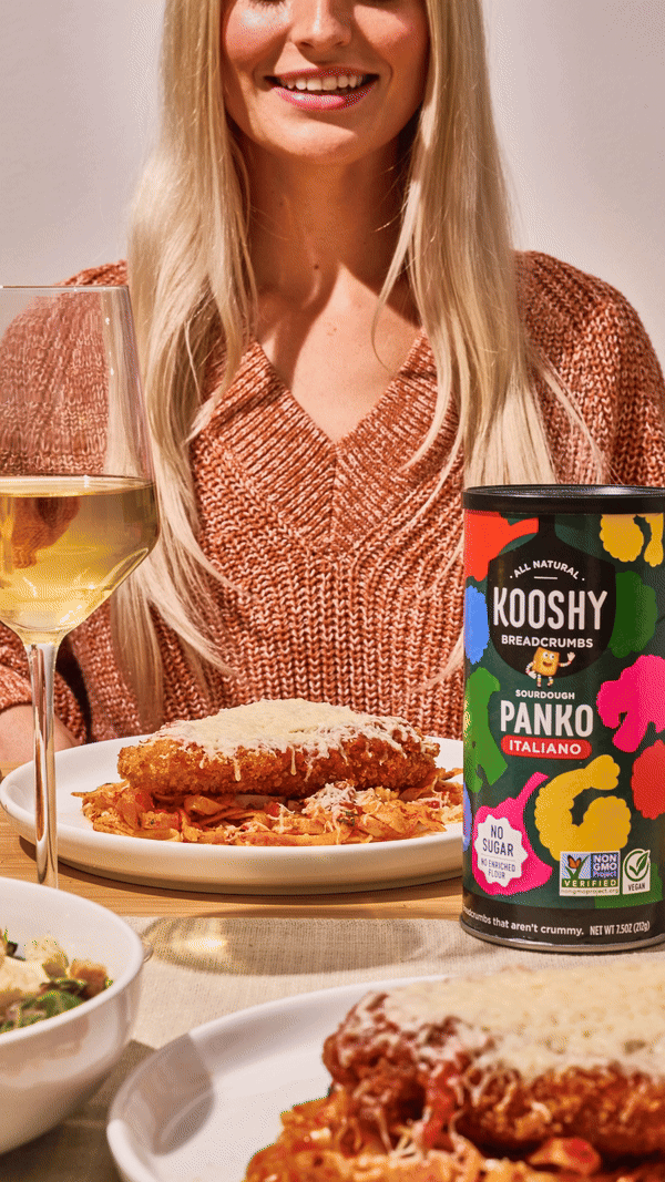 woman eating chicken parm with a knife and fork on a dinner table with a glass of wine and can of kooshy panko breadcrumbs.