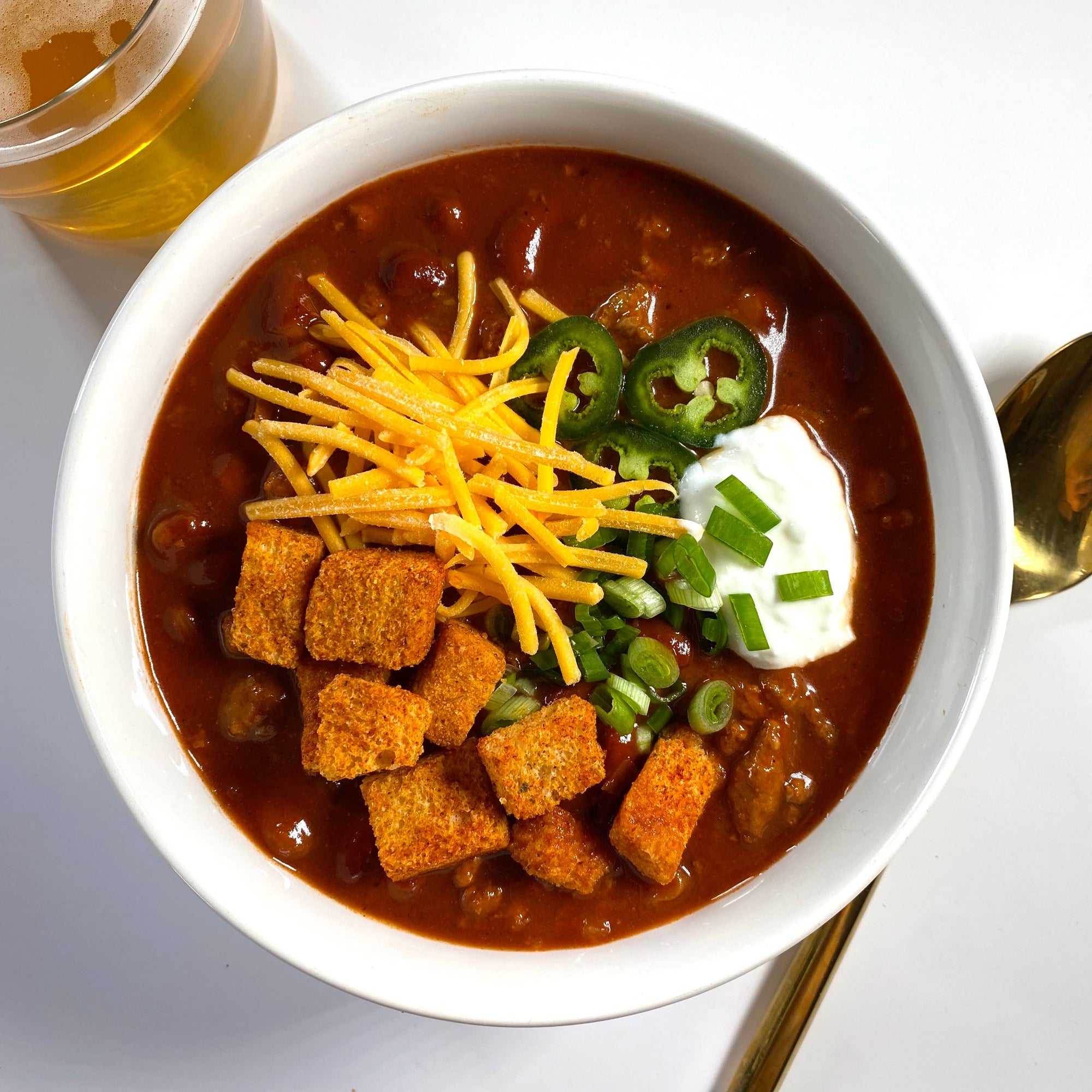 Bowl of chili with croutons, jalapenos, sour cream, green onion and cheddar cheese next to a glass of beer on a white table.