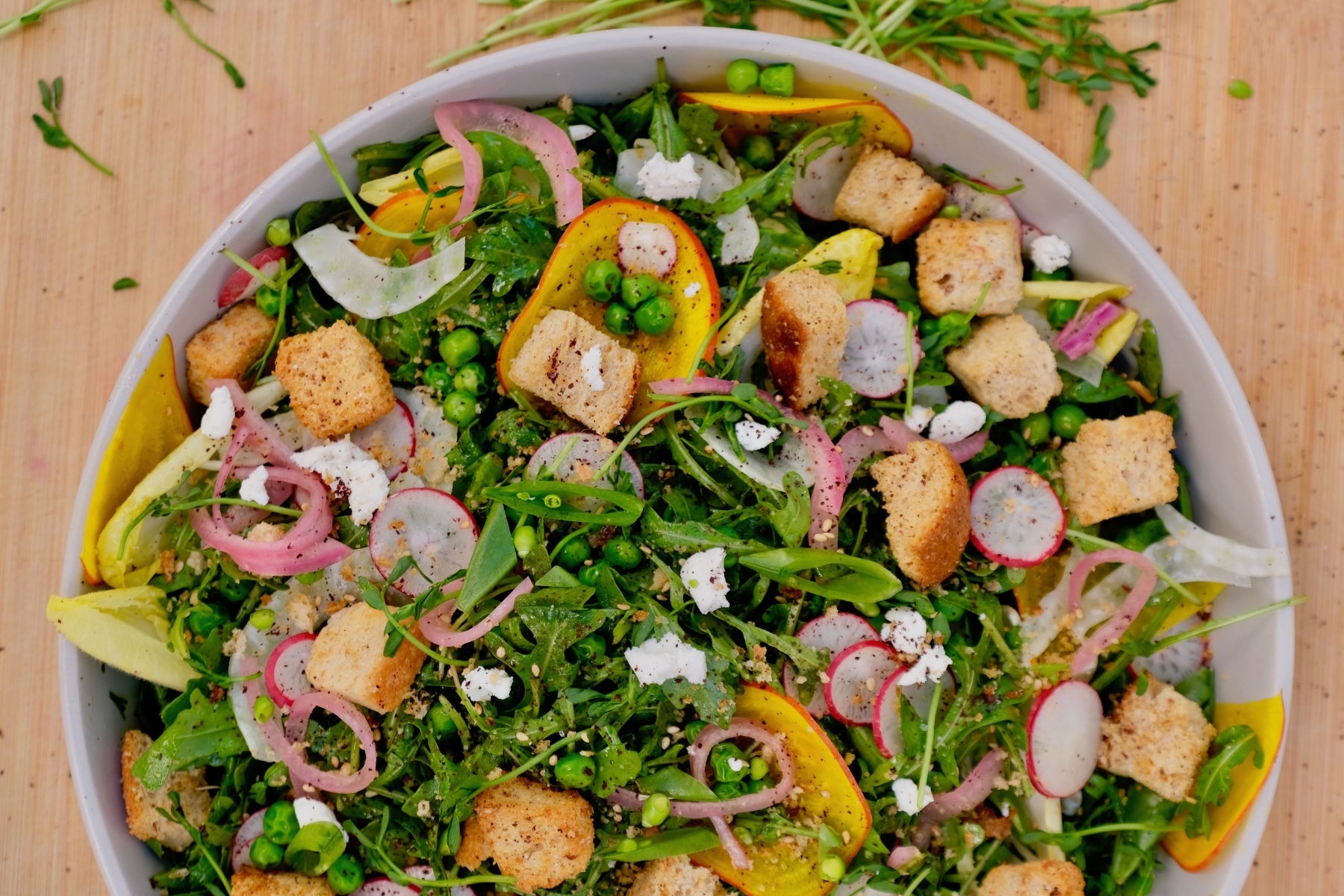 a colorful spring salad in a bowl containing croutons, raw beets, onions, fennel, feta cheese and peas all on top of a wooden counter.