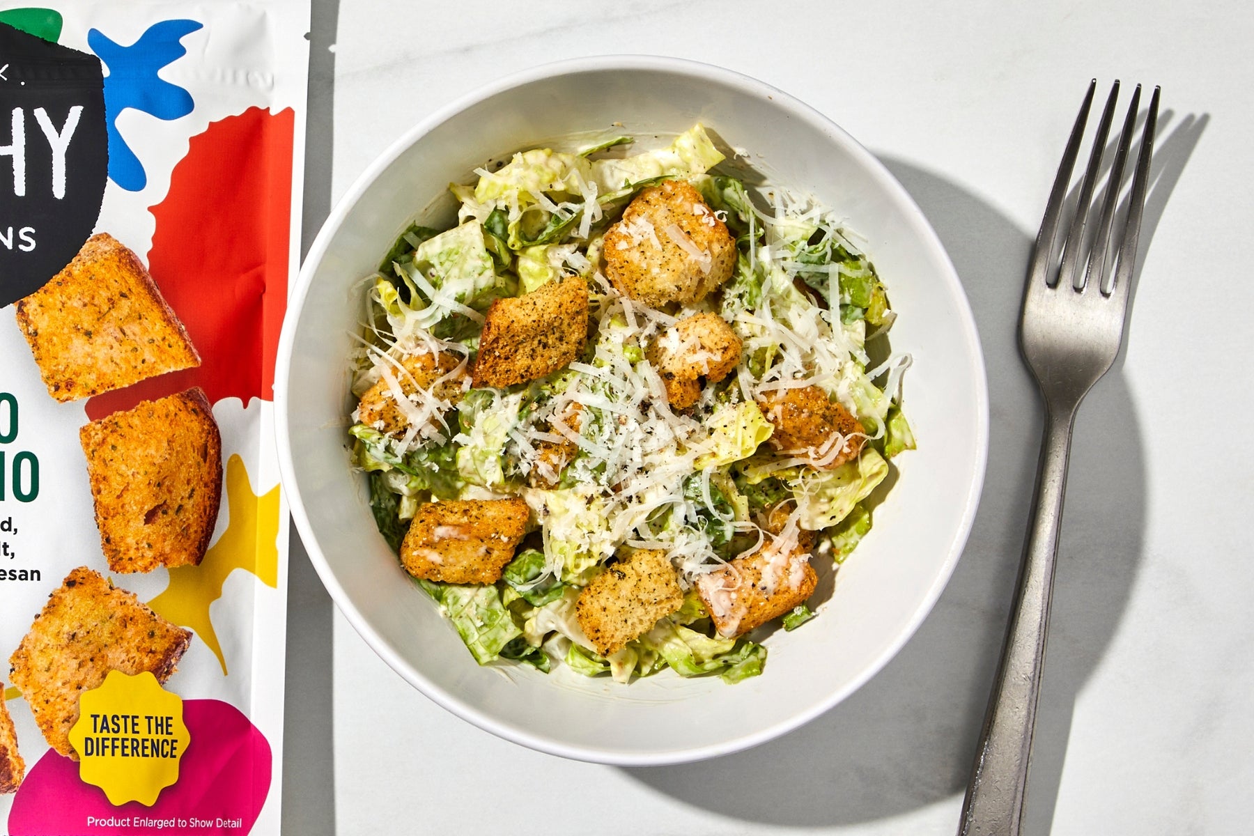 a bowl of caesar salad with croutons, a silver fork and a bag of mambo italiano croutons.