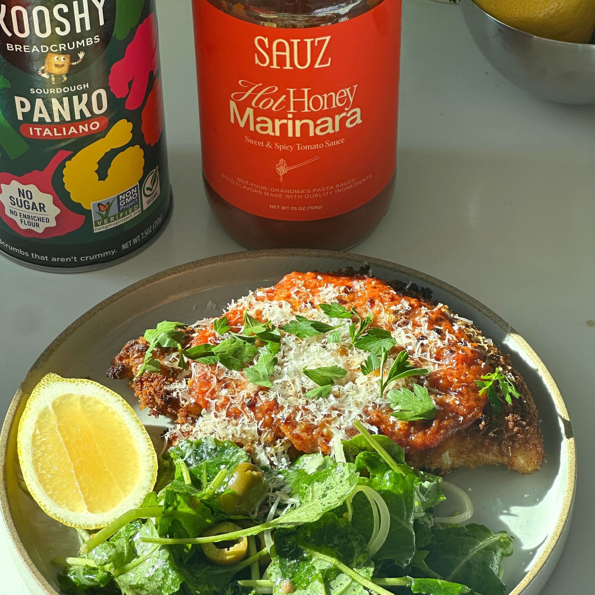 a plate of chicken parm with a side salad with a lemon wedge and packages of panko breadcrumbs and tomato sauce behind them.