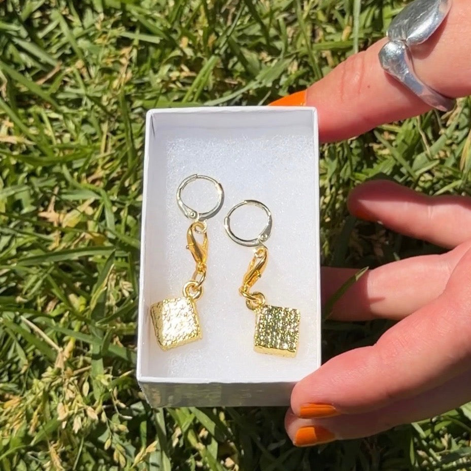 Gold book-shaped earrings in a box held by a hand with orange nail polish against a grass background.
