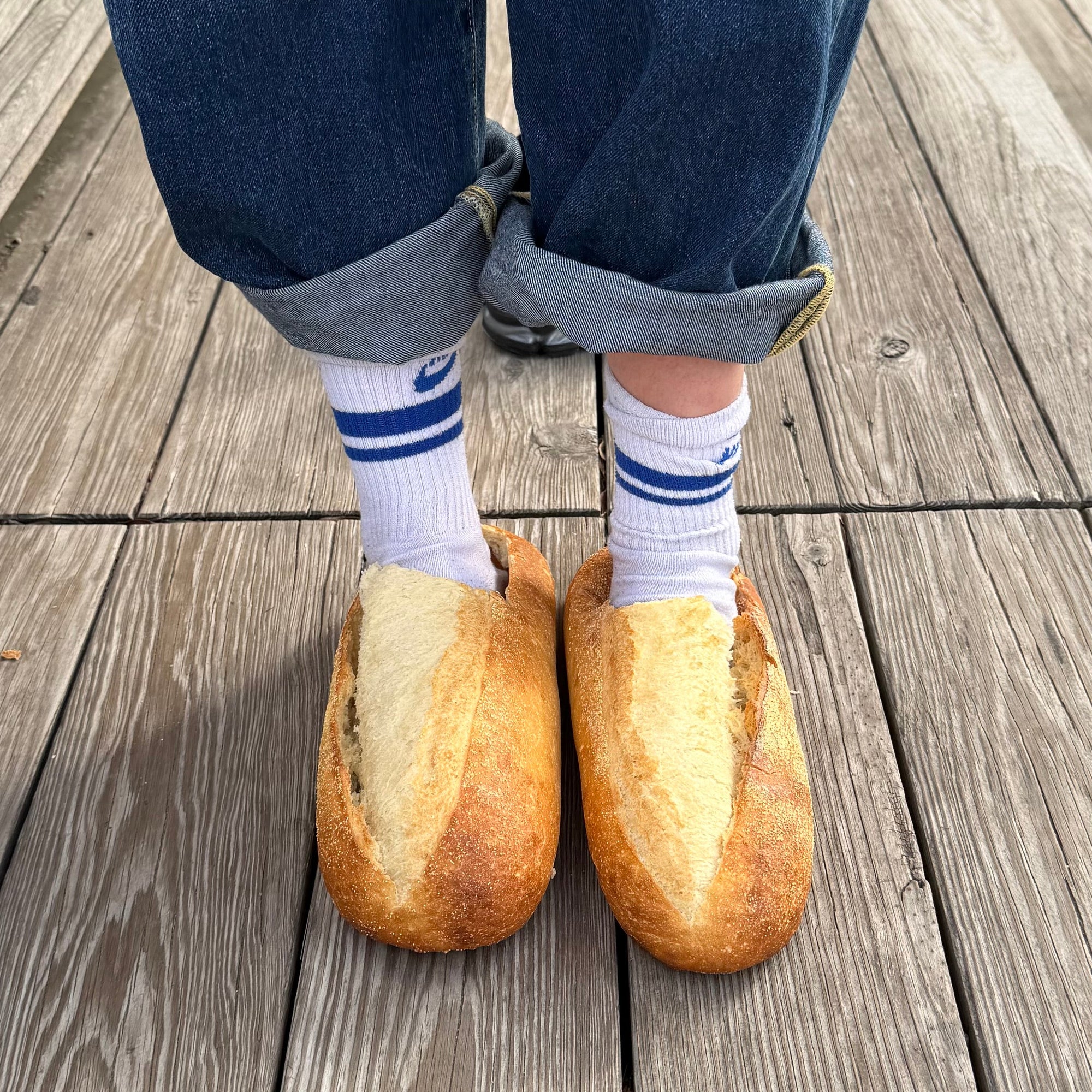 Bread-shaped slippers worn on a wooden floor with rolled-up jeans and striped socks.