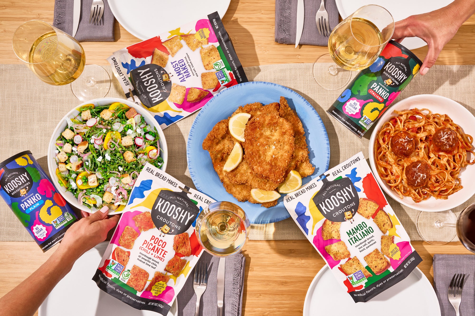 Overhead image of a dinner table with packages of kooshy croutons and breadcrumbs alongside a bowl of salad, meatballs and chicken cutlets. 