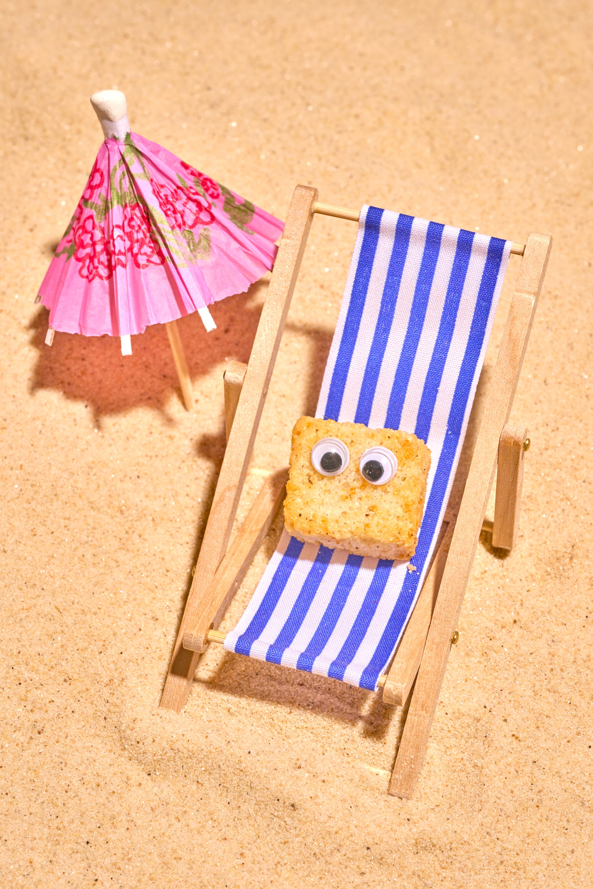 funny photo of a small crouton prop with googly eyes in a miniature beach chair with an umbrella in the sand.