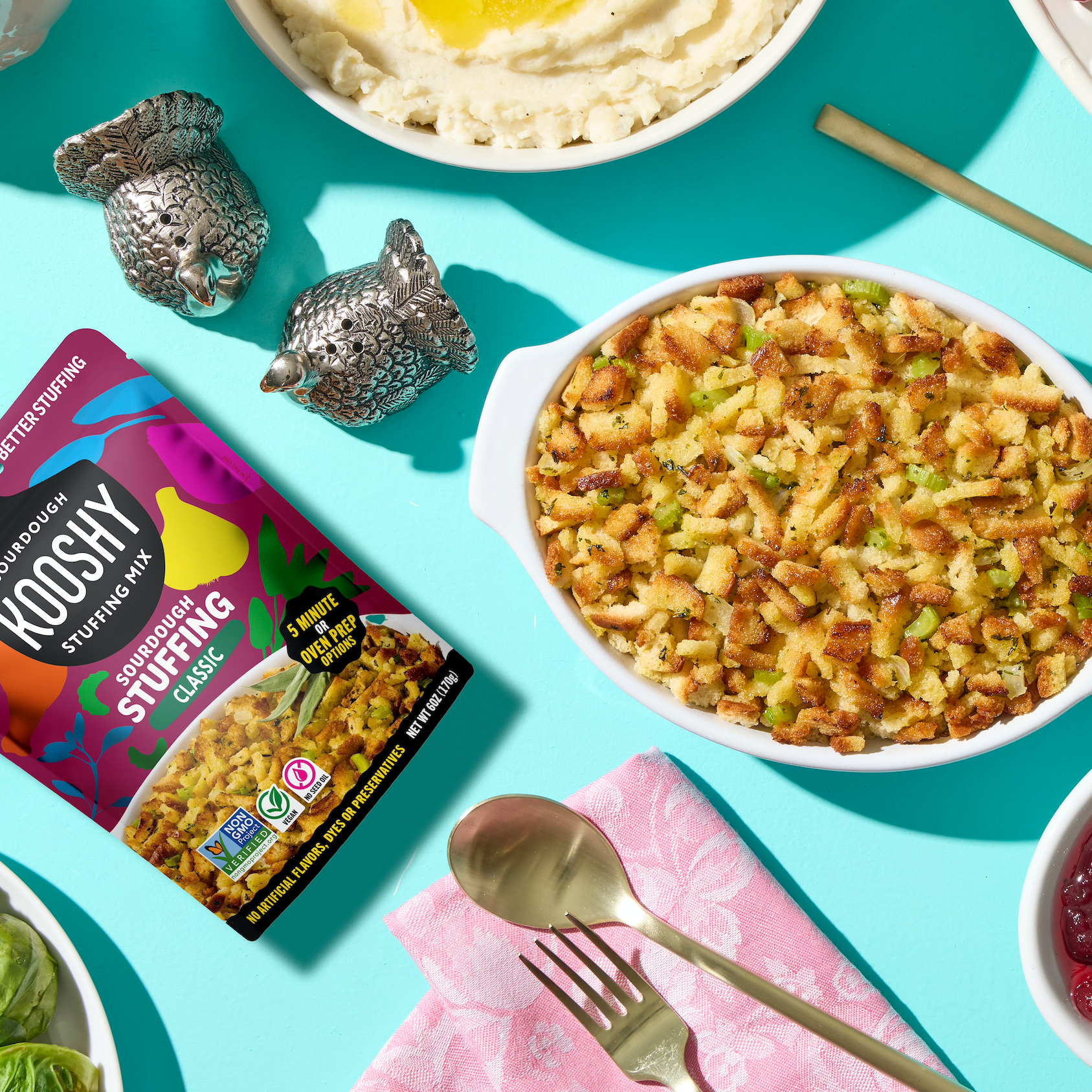 Assorted holiday foods including a bowl of stuffing, cranberry sauce, and a package of Kooshy stuffing on a turquoise background.