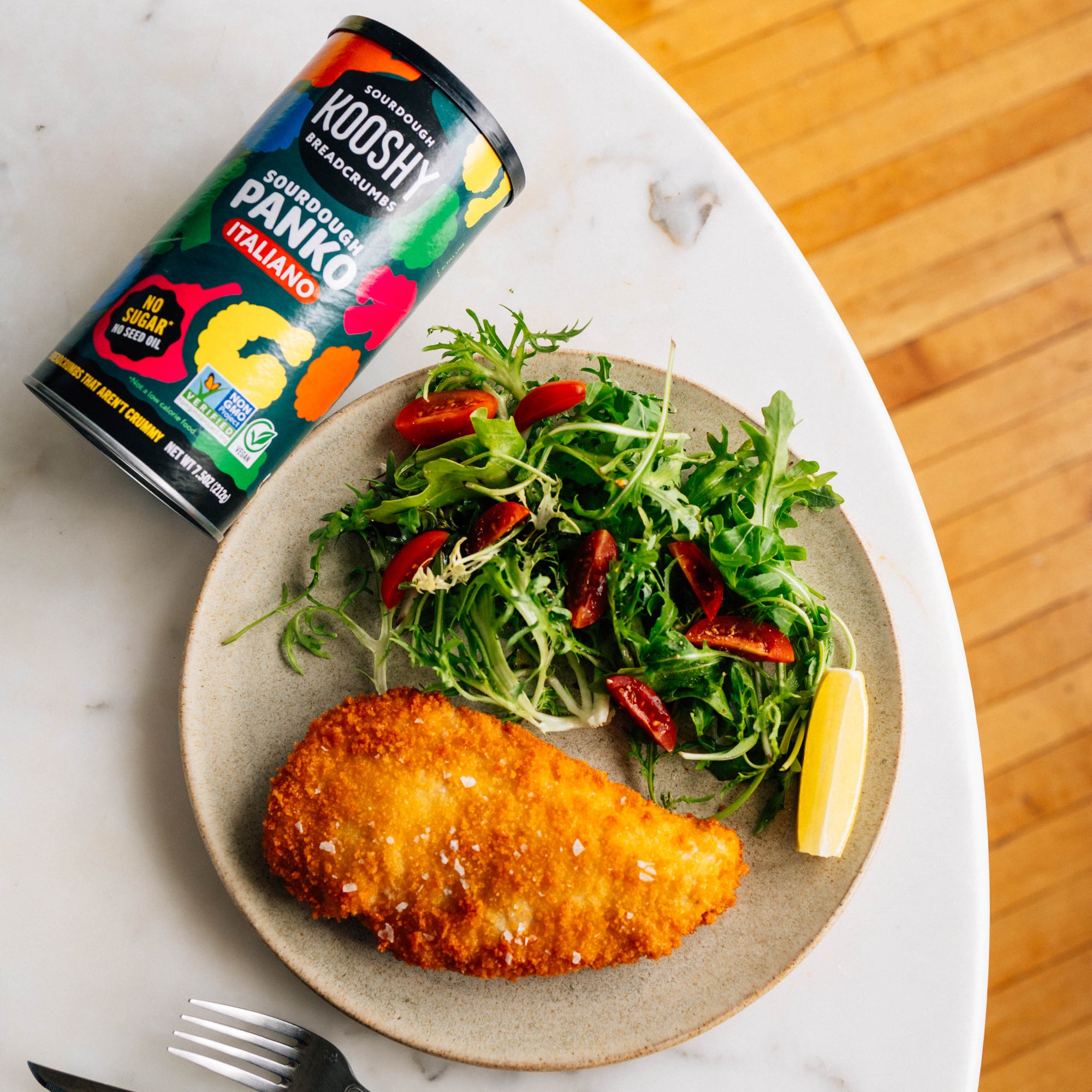 Fried chicken dish with salad on a plate, accompanied by a can of Koshik Panko on a marble surface.